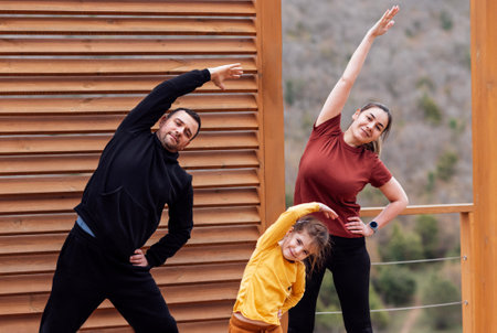 Happy family are stretching outdoors. Attractive mom, handsome dad and cute daughter doing sport exercises. Young couple and their kid are training in the park. Smiling man, woman and girl go in for sports.の写真素材