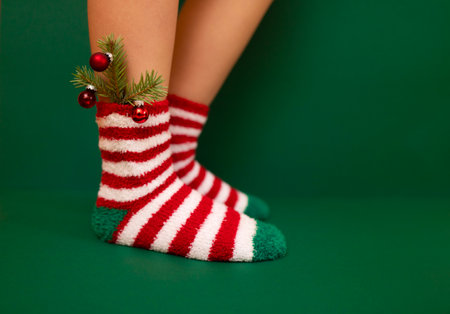 Gils feet in fluffy New Year warm socks. Christmas tree branch with balls decorats female leg. The colors of the socks are red and white stripes and green heels and tips. green background.の写真素材