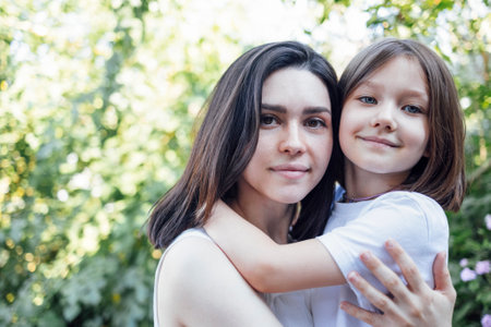 Young charming woman is hugging her cute little daughter outdoors in garden among green trees. Happy mom and kid is cuddling and having a good time together in the park. Mothers day and childhood concept.の写真素材
