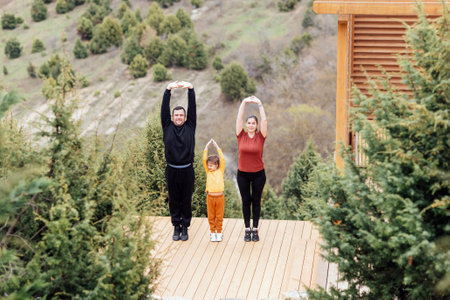 Happy family are stretching outdoors. Attractive mom, handsome dad and cute daughter doing sport exercises. Young couple and their kid are training in the park. Smiling man, woman and girl go in for sports.の写真素材