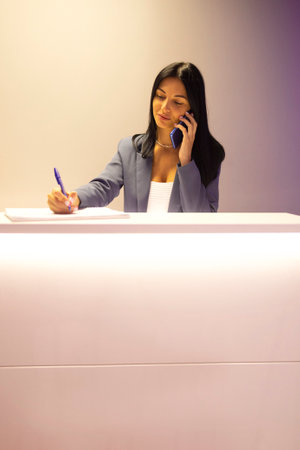 Young beautiful focused receptionist speaks on phone and writes down notes about client. Attractive brown-haired girl stands at reception and makes a call. Product mockups on table. Modern interior.の写真素材