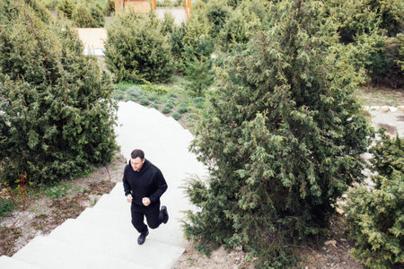 Young man is jogging in the springtime. Active male athlete in black sportswear running up the stairs. Sportsman training early in the morning through the park. Top view. Healthy lifestyle concept.の写真素材