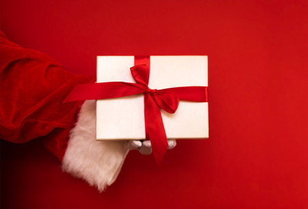 Close up of a hand of Santa Claus in white glove holding a present box. Arm with white wrapping paper gift and red bow. Bright red background. Christmas holiday celebrate concept. Happy New Year eve.の写真素材