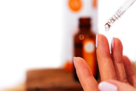 Extreme close up of womans hand and pipette. Transperent drop of essence, oil or gel drips from the pipette. Blurred brown glass serum bottle on the background. Concept of natural organic cosmetic.の写真素材