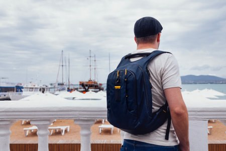 A young man in gray shirt and jeans with a black backpack on the seashore. A male in a cap enjoys...