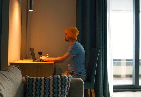 Young man in casual clothes and yellow hat sits with laptop and wine glass in cozy living room. Male...
