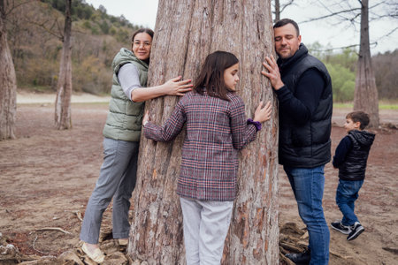 Young attractive woman, man and pretty teenage girl are hugging large cypress trunk. Happy family walks among trees. Little cute boy looks around. Protecting environment and caring for plants concept.の写真素材