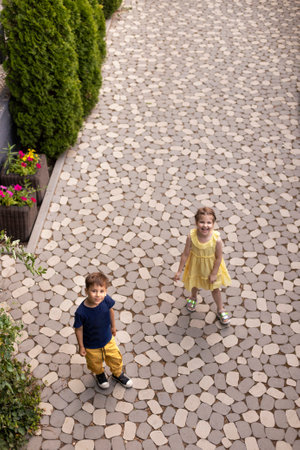 Little funny children laugh and look at the camera. Cute boy and girl in casual clothes have fun in the park. View from above. Stone path, arborvitae and flowers on the background.の写真素材