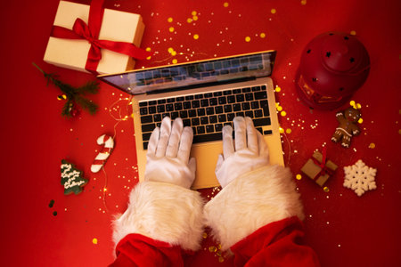 Santa Claus with laptop. Hands in white gloves typing on an notebook keyboard. Gift boxes, christmas cookies, lantern and fir branch on red background. Bright backdrop with sparkles and confetti.の写真素材