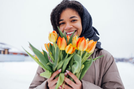 Young african woman in warm casual clothes holds bouquet of orange tulips. Joyful attractive afro...