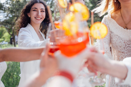 Close up of young women holding elegant glasses with long stem of alcohol cocktails and clink them. Making a celebratory toast with delicious citrus drinks with slices of orange. Party outdoors.の写真素材