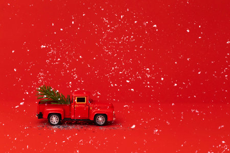 Red toy truck with Christmas fir branch in its trunk. Miniature car with Christmas decoration on red background. Bright backgdrop with sparkles and confetti. New Year holiday concept.の写真素材