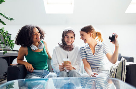 Three pretty female teenagers of different nationalities are having fun and laughing. Caucasian, African and Muslim teens girls take selfie on vintage camera at home. Modern interior with skylights.の写真素材