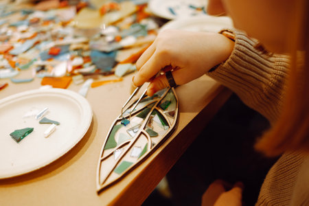 Female hand with rings holding glass piece with tweeters while making mosaic. Teen girl inserts green piece of glass into shape of tree leaf. Multicolored pieces of glass and ceramics on background.の写真素材