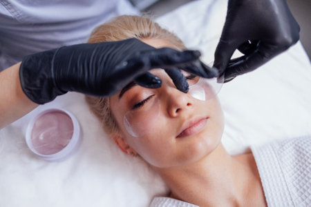 Procedures in a beauty salon. Close-up of a master applying silicone patches under the eyes of a pretty female blond client. Caucasian young woman gets professional facial skin care.の写真素材