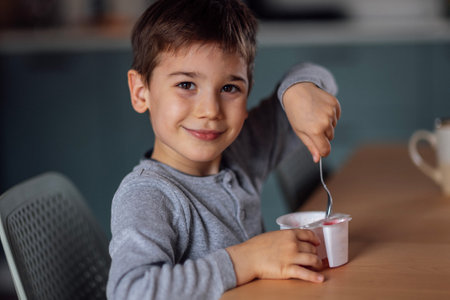 Close up of portrait of cute little boy eats delicious yogurt or cottage cheese from white container with spoon at table in dining room. Happy child smiles and has his breakfast at home. Kid morning.の写真素材