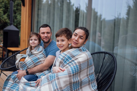 Happy family with two children is sitting on the terrace at home. Young parents covered their little kids with checkered blankets. Laughing dad hugging cute daughter. Beautiful nature on background.の写真素材
