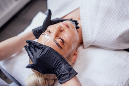 Close up cosmetologist hands in black gloves apply transparent vitamin mask to her client face in spa salon or clinic. Blonde beautiful girl in white bathrobe is undergoing skin moisturizing procedure.の写真素材