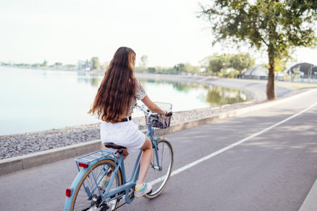 Back view of female teenage is driving her bicycle. Young and positive girl in casual clothes has a fun. City street and trees with bushes in the background. copyspace.の写真素材