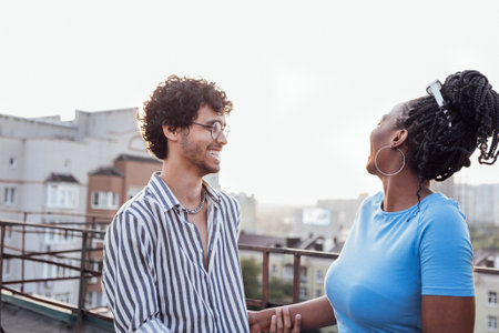 Young happy couple in love on the roof of multi-storey building. Cute curly-haired interracial guy holds hand of charming African woman. Multiethnic smiling friends against backdrop of urban landscape.の写真素材