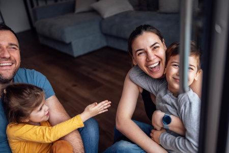 Young parents and small children sit on floor of living room at home in front of open panoramic window. Mom and dad hug and wrap their kids in checkered blankets. Tender moment family care and love.の写真素材