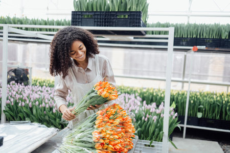 Cute attractive african girl in green apron holding bouquet of orange tulips indoors in greenhouse. Female darkskinned flower farm employee is packing flowers for delivery in shops. Copy spaceの写真素材