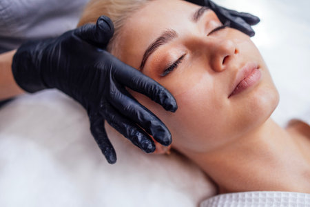 Procedures in a beauty salon. Close-up of a master applying silicone patches under the eyes of a pretty female blond client. Caucasian young woman gets professional facial skin care.の写真素材