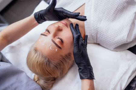Close up of portrait of a young attractive woman getting facial massage with cream. The blonde girl enjoys beauty procedure. Masseuse hands in black gloves on the female client chin. Spa treatment.の写真素材