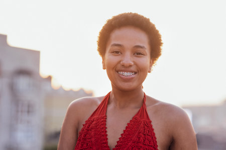 Close-up of female portrait of young adult attractive confident African. Charming darkskinned girl in red top stands against background of big city. Mixed race woman looks into camera and smiles.の写真素材