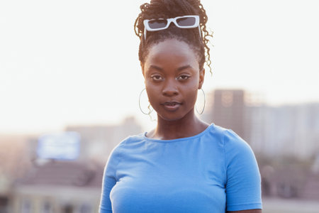Close-up of female portrait of young beautiful African. Charming black girl with sunglasses and blue T-shirt stands against background of city. Mixed race woman looks at camera and smiles.の写真素材