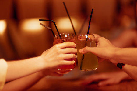 Teenagers raise a festive toast with cocktails in glasses with straws. Friends have a good time in a cafe with a modern interior. Guys and girls are sitting at a table in a restaurant and clinking drinks.の写真素材