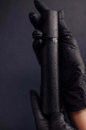 A mockup of a black tube for cream with drops of water. Close-up of the beautician hands in black gloves holding a moisturizer on a dark isolated background. Advertising cosmetics.の写真素材