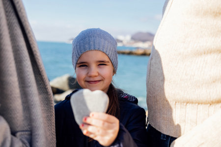 Cute little girl stands between her parents and shows a heart-shaped stone. A sweet baby in a...