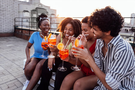 Company of four cheerful friends are enjoying tasty cocktails. Roof party. Interracial man and African women drink aperol with orange slices. Multiethnic group of people have fun and laugh together.の写真素材