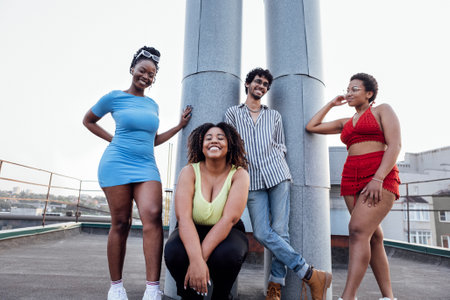Company of four multi-ethnic people pose on the roof of multi-storey house. Stylish portrait of friends of mixed race against background of metal pipes and city. Guy and girls laugh and have fun together.の写真素材