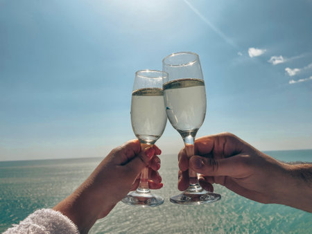 Close-up of two clinking glasses of champagne against the blue sea and sky. Female and male hands holding goblets with sparkling wine and making a toast. Vacation, date celebration, holiday concept.の写真素材