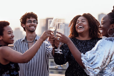 Dark-skinned girl pours champagne into glasses of her multinational friends. Cute happy people having fun at rooftop party. African women in elegant dresses and young man drink wine and laugh.の写真素材