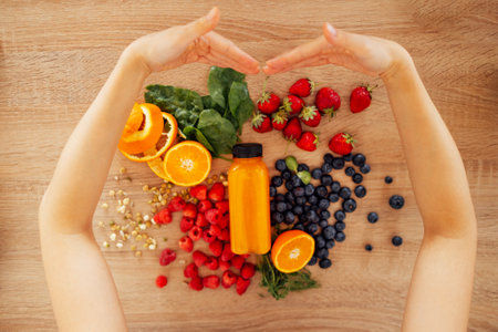 Female hands embracing citrus fruits, berries, greens, sprouted beans and bottle of orange juice on the background of a wooden table. Heart gesture. View from above. Healthy food and drinks concept.の写真素材