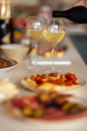 Two glasses of cool champagne and plates with various snacks on a table. White plates with slices of ham, salami, capers, olives and cheese. Cozy interior of the living room of the house.の写真素材