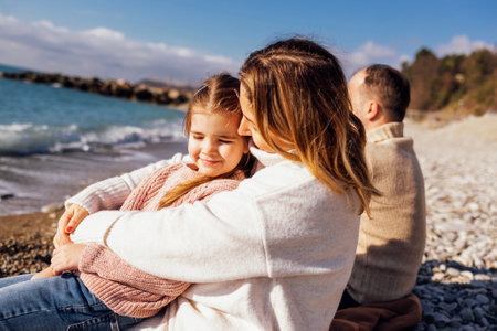 Man, woman and little child by the sea. Mom, dad and female sitting back. Cute little girl smiling and looking at camera. People are dressed in warm sweaters. Happy family have weekendの写真素材