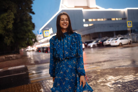 Beautiful brunette girl in blue dress laughs and dances in pouring rain in evening. Young beautiful woman is having fun on city street in bad weather. Parking cars and shopping mall on background.の写真素材