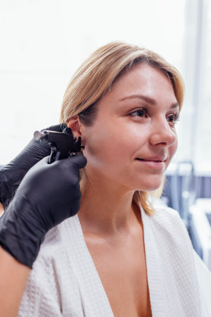 Close up of face of young blonde woman doing ear piercing procedure. Doctor hands in black gloves...