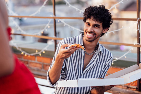 A cute mixed-race guy smiles and holds a slice of pizza in his hand. A young latin man with curly hair laughs in a striped shirt and has a snack. Roof party. Garland, fencing and houses on background.の写真素材