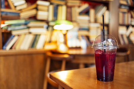A red lemon cocktail with ice cubes and a straw stands on a wooden table in the library. Large...