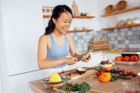 An attractive cute Asian woman smiles and cooks breakfast in the kitchen. A young charming Korean woman laughs and cuts vegetables and fish. Healthy food. Cozy modern interior of the room.の写真素材