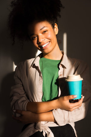 An attractive young girl of mixed race holds a cup of coffee and smiles. A charming multiethnic teenager in casual clothes is drinking tea to take away. Beautiful African woman with a hot drink.の写真素材