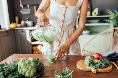 A happy curly-haired girl in a linen sundress is preparing a healthy breakfast. A young smiling woman holds a blender with a green smoothie. Cabbage, broccoli, arugula are on the table.の写真素材