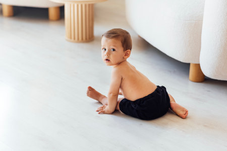 A cute baby is sitting on the white wooden floor in the living room. The little boy toddler is playing at home. The sweet child looks at the camera with interest. Copy space.の写真素材