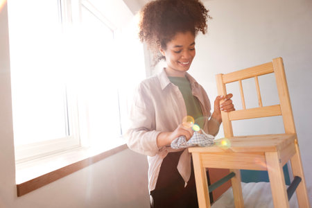 A beautiful smiling mixed-race girl wipes a wooden chair with a rag. An attractive young African woman is preparing furniture for painting. Renovation in an apartment or house.の写真素材