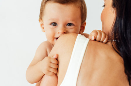 A young woman holds her beautiful baby. Charming mom with a sweet cute toddler on a white wall...